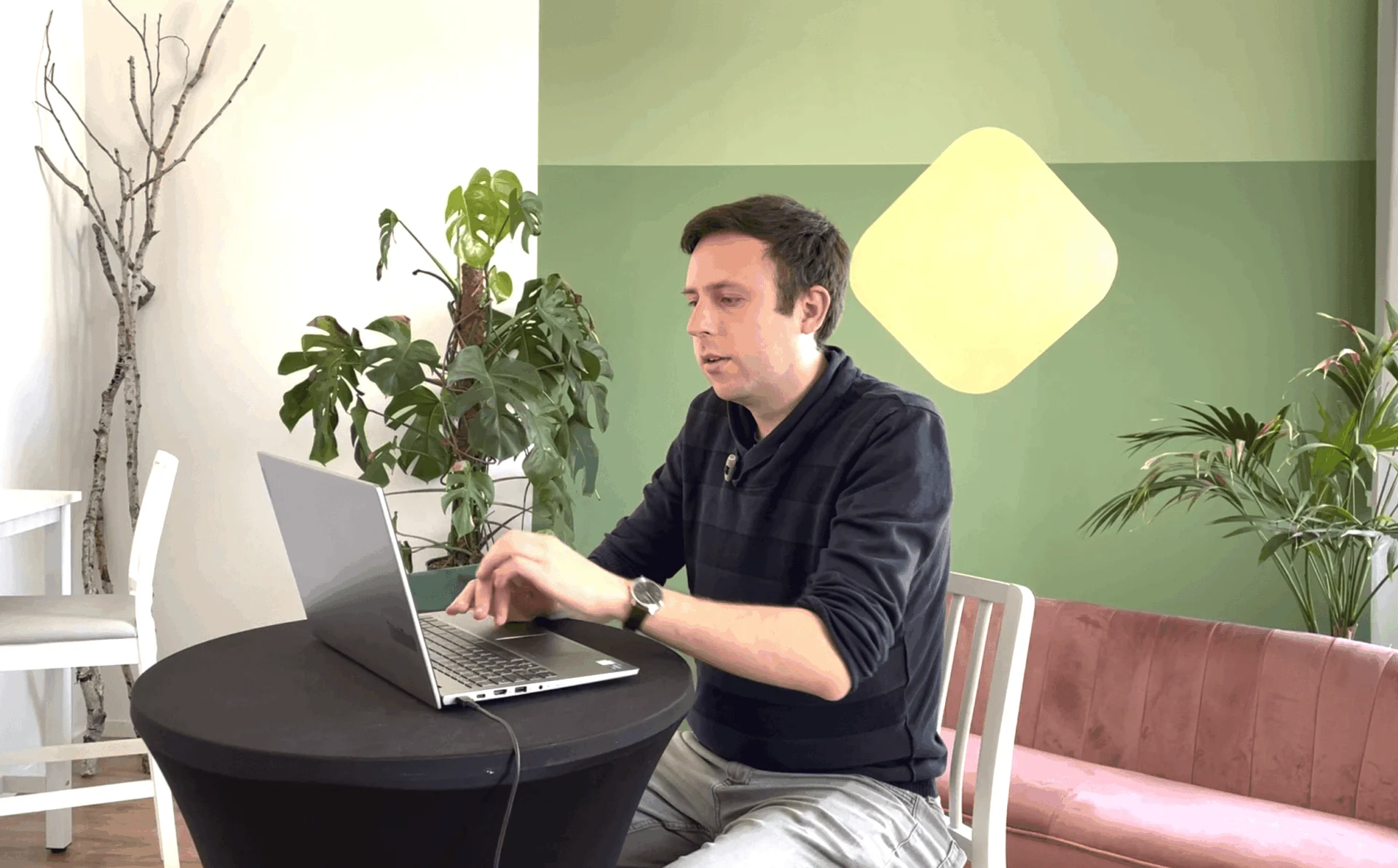 Ein Mann sitzt an einem runden schwarzen Tisch und arbeitet an einem Laptop, im Hintergrund stehen grüne Pflanzen und ein rosa Sofa.
