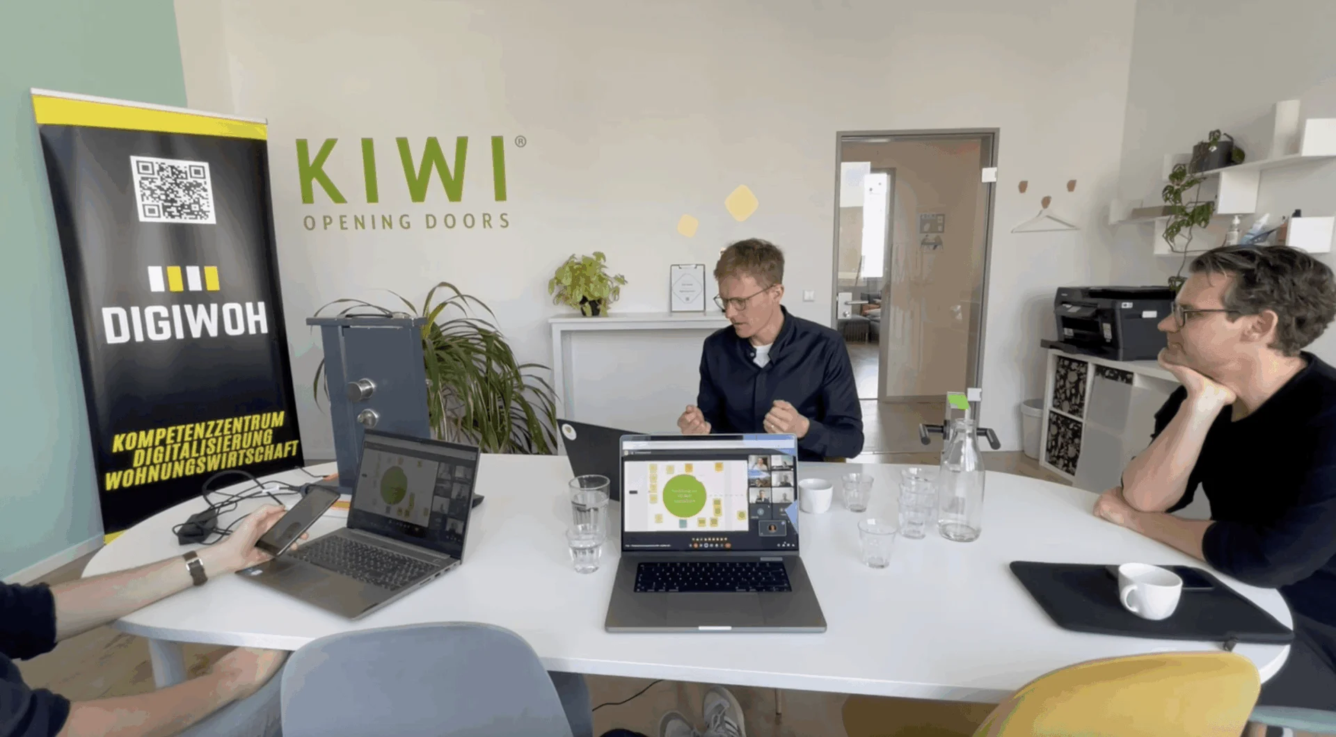 Drei Personen sitzen mit Laptops und Brillen um einen Konferenztisch und nehmen an einer Besprechung in einem modernen Büro teil, an dessen Wand die Banner "KIWI OPENING DOORS" und "DIGIWOH" hängen.