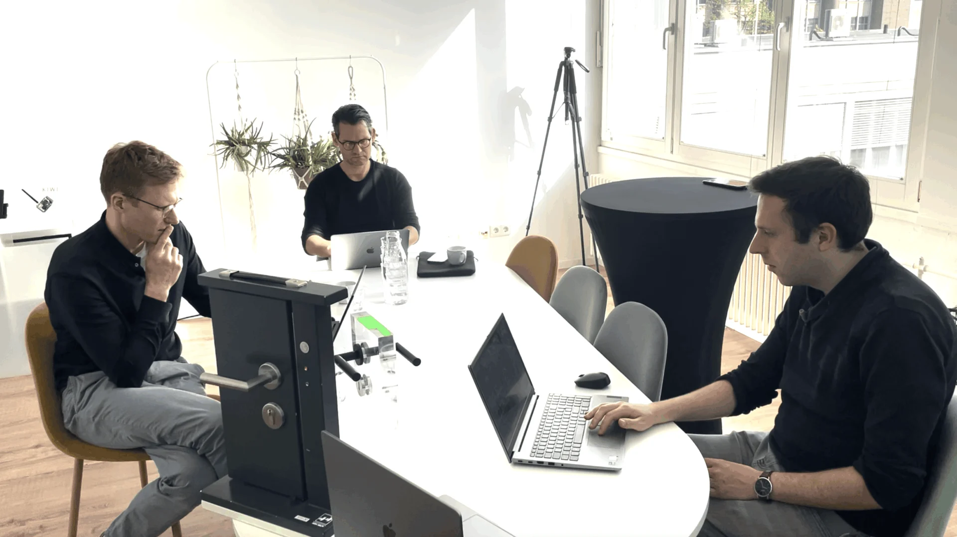 Drei Männer sitzen mit Laptops an einem Konferenztisch und arbeiten in einem hellen Büro mit großen Fenstern und einer Pflanze im Hintergrund.