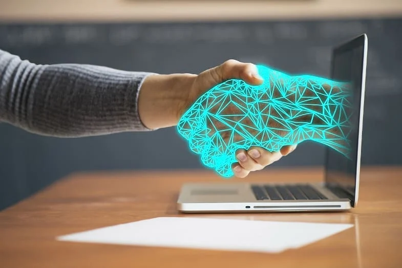 Eine Person schüttelt einer digitalen Hand die Hand, die aus einem Laptop-Bildschirm herausragt und Technologie oder virtuelle Zusammenarbeit symbolisiert.