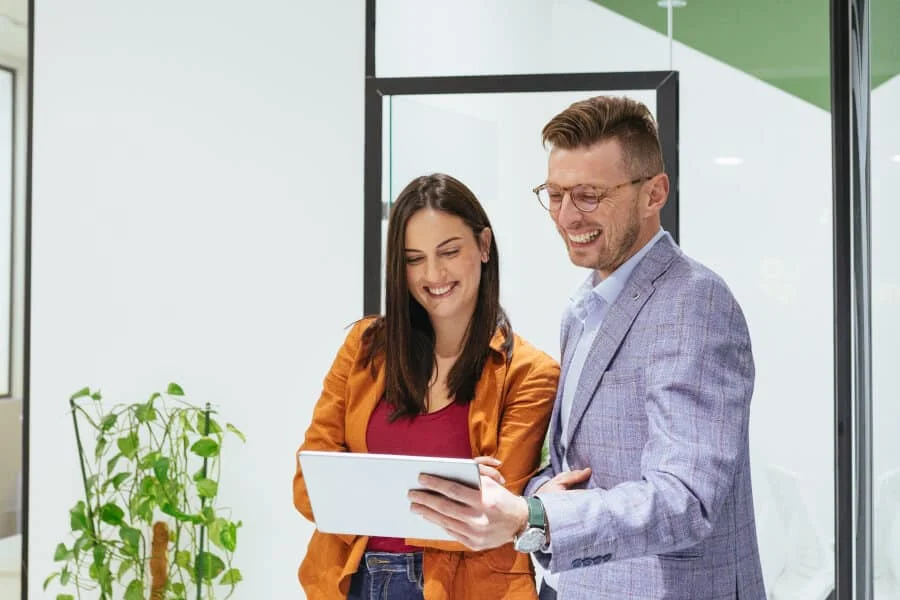 Zwei Personen stehen in einem Büro, lächeln und schauen gemeinsam auf ein Tablet. Im Hintergrund ist eine Grünpflanze zu sehen.