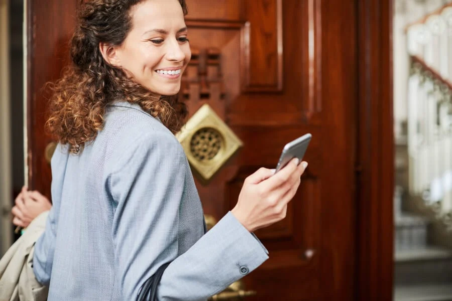 Eine Frau in einem Blazer, lächelnd und mit einem Telefon in der Hand, steht vor einer Holztür mit Messingbeschlägen, die sie gerade betreten oder verlassen will.