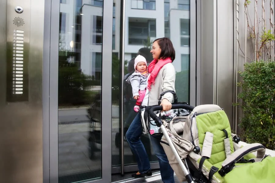 Eine Frau mit einem Baby im Arm und einem leeren Kinderwagen betritt ein modernes Gebäude durch eine Glastür.