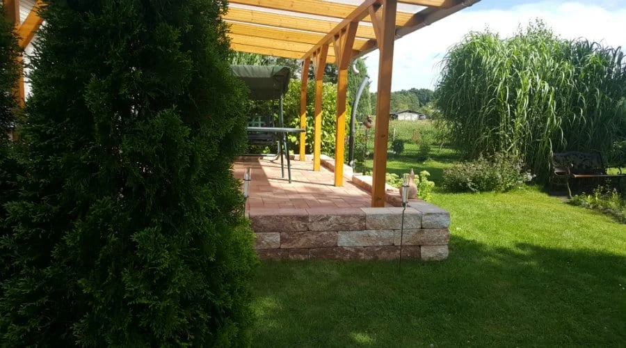 Eine hölzerne Pergola mit einem Steinsockel steht auf einer Terrasse in einem grünen Garten mit Rasen, Sträuchern und Gartenmöbeln unter einem teilweise bewölkten Himmel.