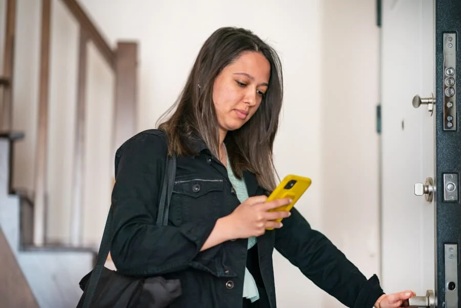 Eine Frau in schwarzer Jacke schließt eine Tür auf, während sie auf ihr gelbes Smartphone schaut und eine Umhängetasche über dem Arm trägt.