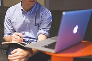 Eine Person in einem Button-up-Hemd sitzt auf einer Couch und macht sich Notizen in einem Notizbuch, während sie auf einen offenen Laptop auf einem kleinen orangefarbenen Tisch schaut.