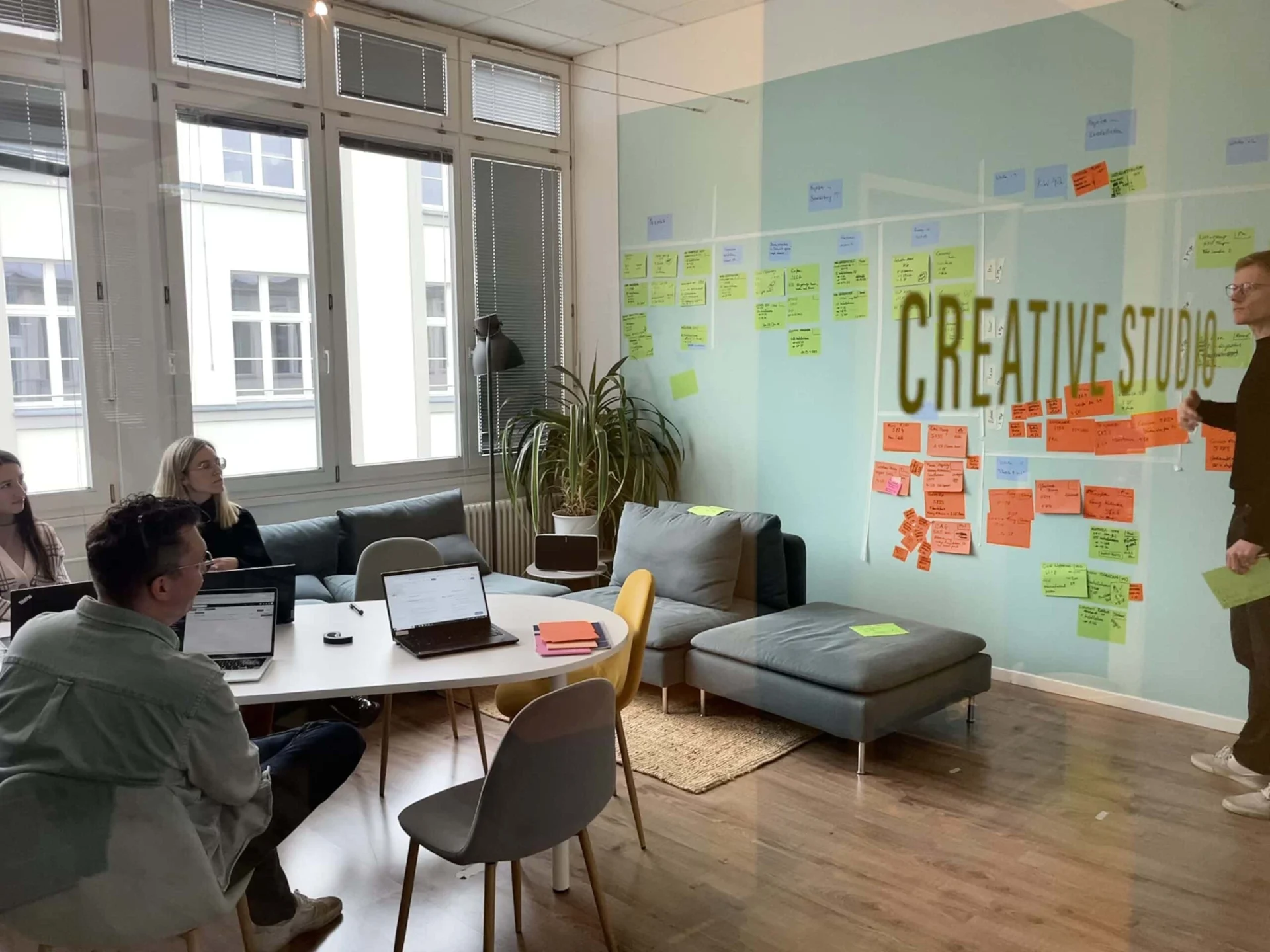 Vier Personen in einem modernen Büro mit Laptops, die Ideen diskutieren; bunte Haftnotizen bedecken eine Glaswand mit der Aufschrift "CREATIVE STUDIO".