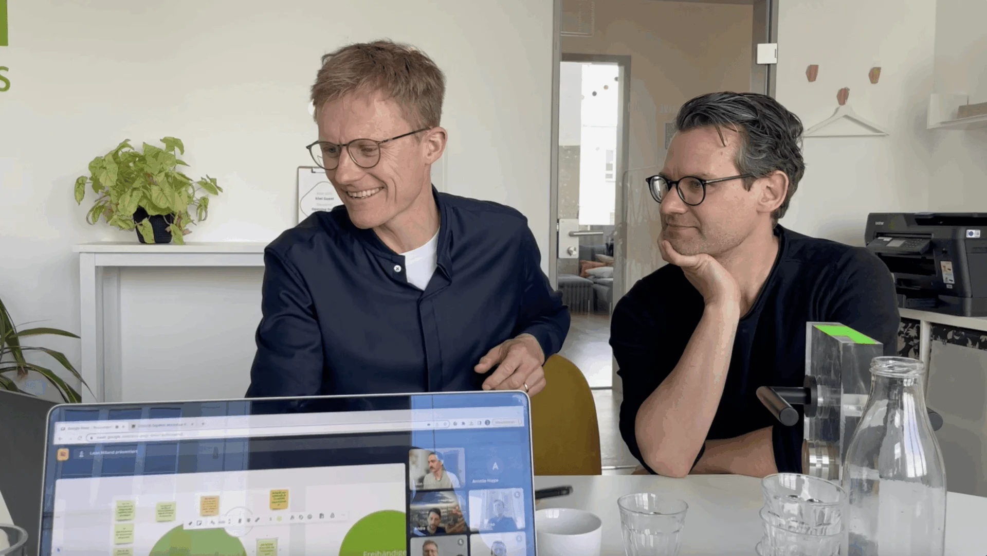 Zwei Männer sitzen mit Laptop, Brille und Tassen an einem Tisch und schauen auf den Bildschirm, während sie an einem Videogespräch in einer modernen Büroumgebung teilnehmen.