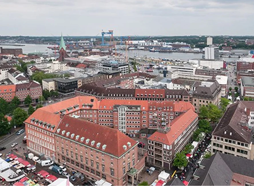 Panoramaaufnahme der Kieler Innenstadt mit Blick auf den Hafen, historische Gebäude mit roten Dächern und Werftkränen im Hintergrund