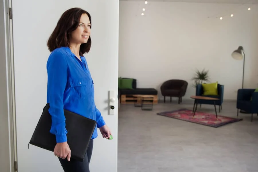 Frau mit blauer Bluse betritt ein modernes Büro, hält eine schwarze Dokumentenmappe in der einen Hand und einen KIWI-Transponder in der anderen.