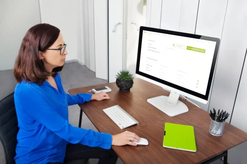 Eine Frau in einem blauen Hemd sitzt an einem Schreibtisch mit einem Desktop-Computer. Auf dem hölzernen Schreibtisch liegen ein Smartphone, ein grünes Notizbuch und eine kleine Topfpflanze.