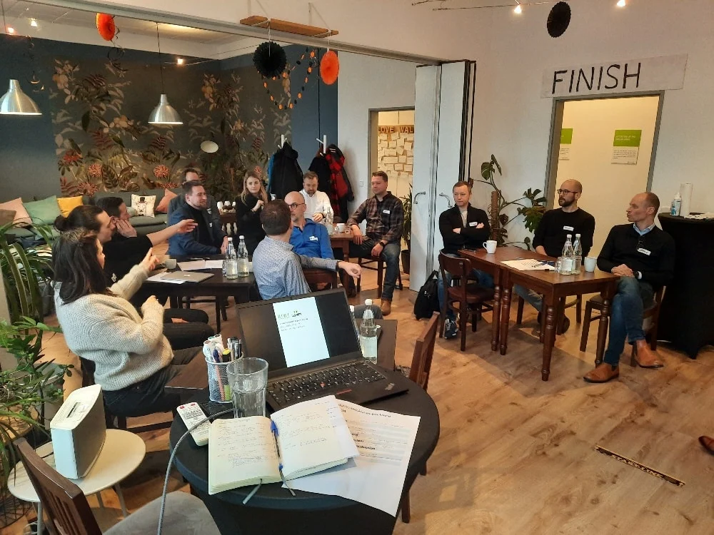 Eine Gruppe von Menschen sitzt in einem gut beleuchteten Raum und diskutiert an Tischen mit Notizbüchern, Getränken und einem Laptop; über einer Tür hängt ein "FINISH"-Banner.