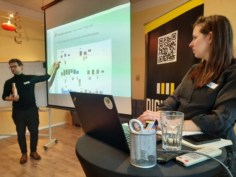 Ein Mann präsentiert ein Diagramm auf einem Bildschirm, während eine Frau an einem Laptop sitzt und Büromaterial, ein Glas Wasser und eine Fernbedienung auf dem Tisch stehen hat. Ein DIG-Banner und ein QR-Code sind zu sehen.