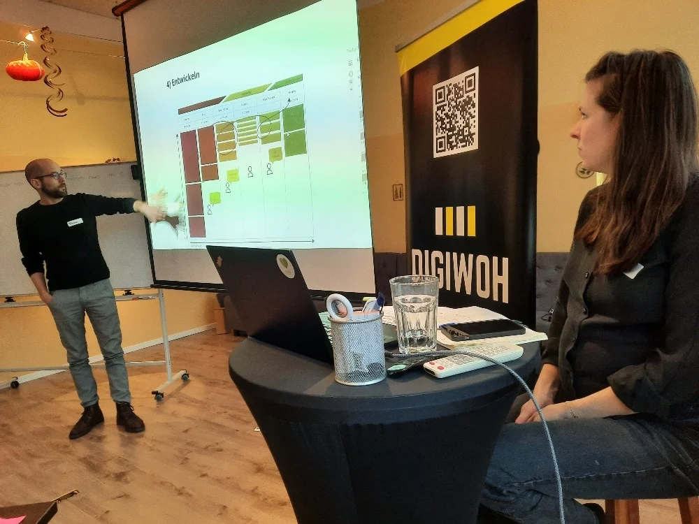 Ein Mann präsentiert einer sitzenden Frau auf einer DigiWOH-Veranstaltung ein Diagramm auf einem Projektorbildschirm, während im Vordergrund ein Laptop und Materialien auf einem Stehtisch stehen.