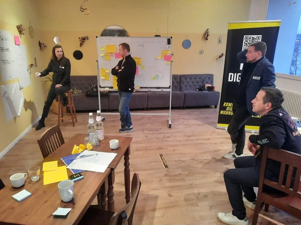 Vier Personen sitzen in einem lockeren Arbeitsbereich und führen ein Brainstorming durch, bei dem Klebezettel auf Whiteboards und Papiere auf Tischen liegen.