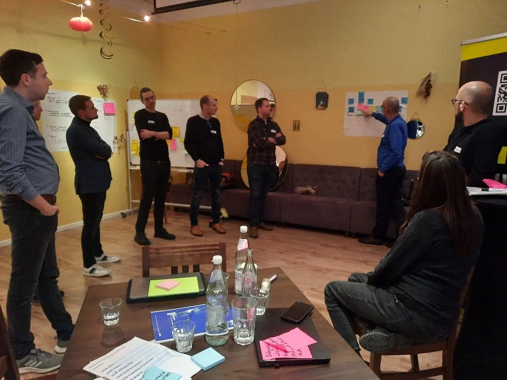 Sieben Personen nehmen an einem Workshop teil; ein Mann steht an einer Tafel mit Haftnotizen, während die anderen beobachten. Auf einem Tisch im Vordergrund befinden sich Notizblöcke, Flaschen und Gläser.