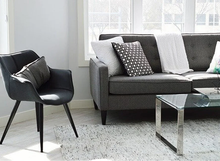 Helles, modern eingerichtetes Wohnzimmer mit grauem Sofa, Designerstuhl, Glastisch und großen Fenstern, im skandinavischen Stil gehalten.