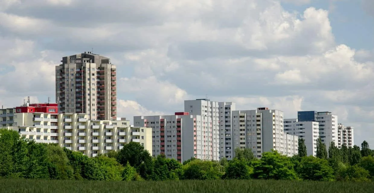 Hinter grünen Bäumen und unter einem wolkenverhangenen Himmel erhebt sich eine Reihe von modernen Wohnhäusern und Hochhäusern.