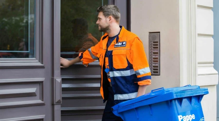 Ein Mann in orangefarbener und marineblauer Uniform steht an einer Tür und klopft, während er eine blaue Recycling-Tonne mit der Aufschrift "Pappe" hält.