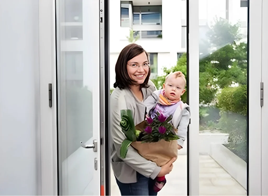 Lächelnde Mutter mit Kleinkind und Blumenstrauß in der Hand betritt durch eine Glastür ein modernes Wohngebäude