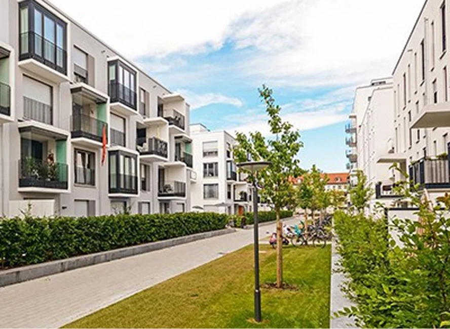 Moderne Neubau-Wohnanlage mit mehreren Mehrfamilienhäusern, Balkonen, Grünflächen und Fußweg unter blauem Himmel.