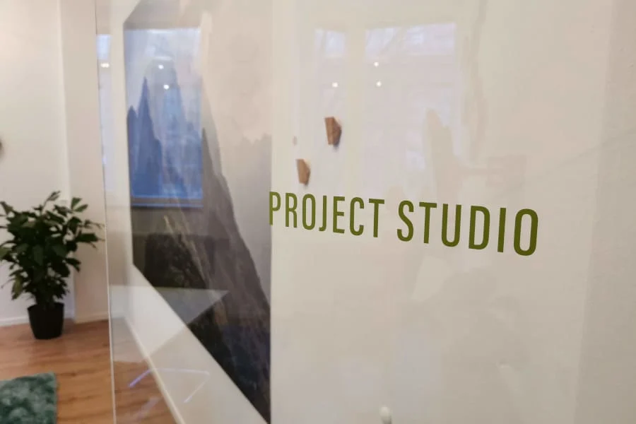 Eine Glaswand mit der Aufschrift "PROJECT STUDIO"; im Hintergrund sind eine Pflanze und ein gerahmtes Kunstwerk zu sehen.