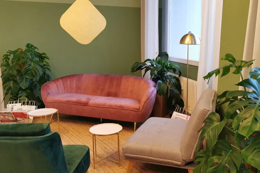 Ein modernes Wohnzimmer mit einem rosa Samtsofa, einem grauen Loungesessel, einem grünen Akzentsessel, kleinen runden Tischen, großen Pflanzen und einem Fenster mit weißen Vorhängen.