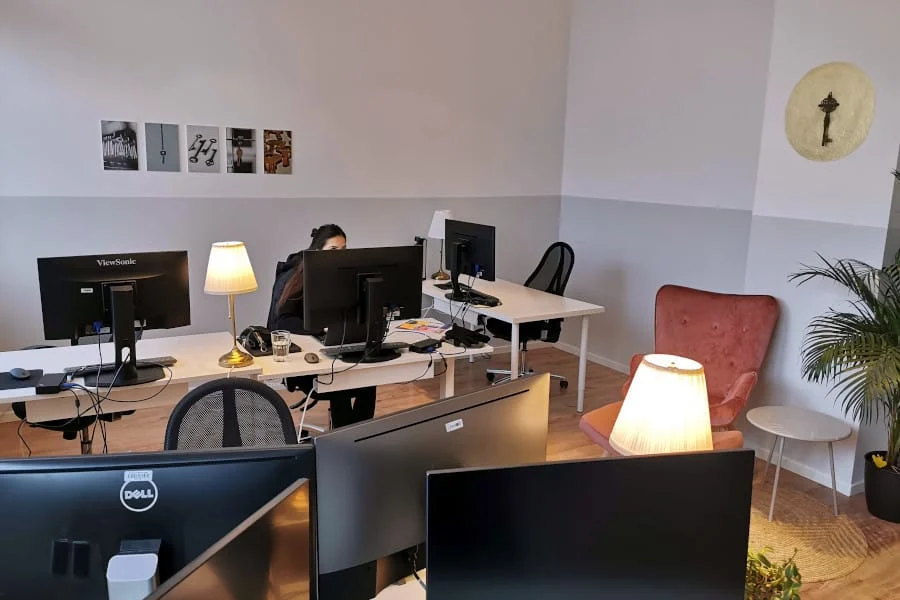 Ein kleines Büro mit mehreren Computertischen, Monitoren, Schreibtischlampen, einer arbeitenden Person, einem leeren Stuhl, Wandkunst und einer Topfpflanze in der Ecke.