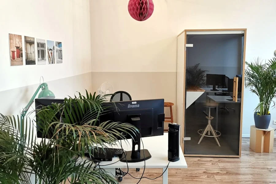 Modernes Büro mit Computern auf Schreibtischen, Zimmerpflanzen, Wandkunst und einer gläsernen Telefonzelle mit einem Hocker und einem Computer darin.