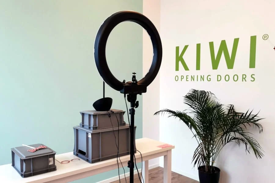 Eine Ringleuchte auf einem Stativ steht auf einem weißen Tisch mit Aufbewahrungsboxen. An der Wand steht "KIWI OPENING DOORS", und in der Ecke steht eine Topfpflanze.