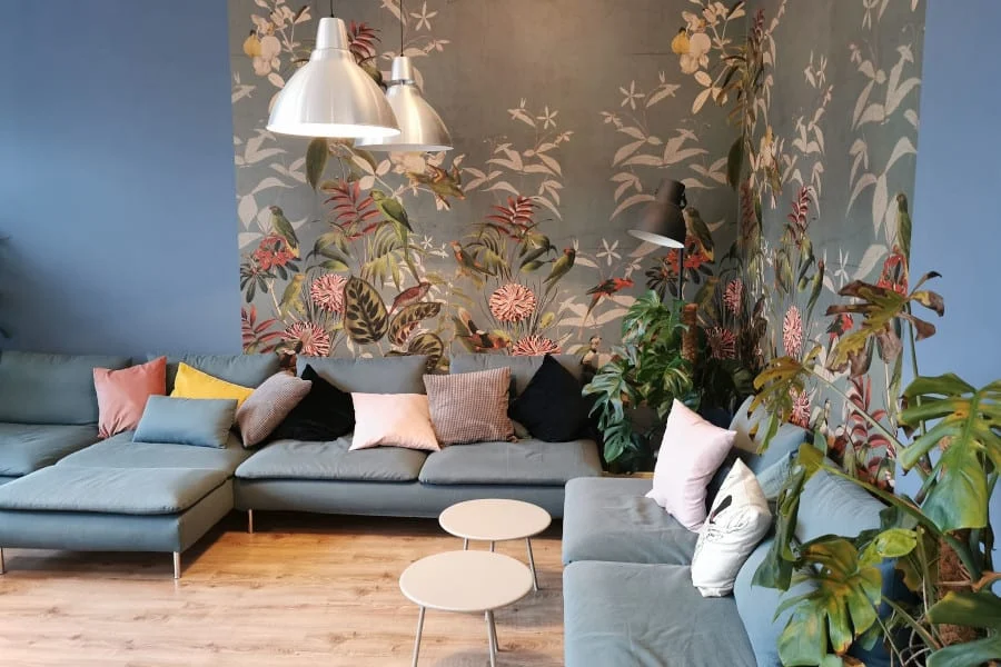 Ein modernes Wohnzimmer mit einem blauen Sektionssofa mit verschiedenen Kissen, kleinen runden Tischen, Blattpflanzen und Blumentapeten an zwei Wänden.