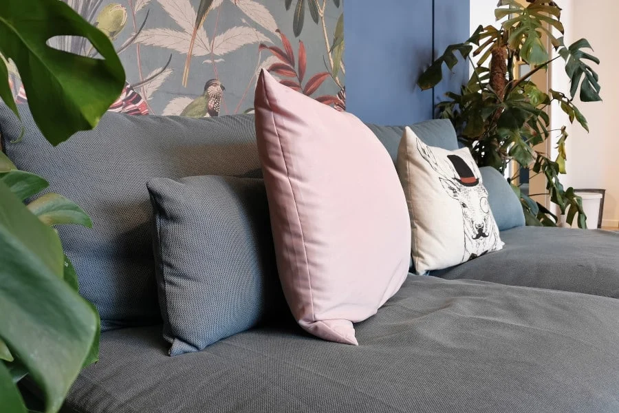 Graues Sofa mit blauen und rosafarbenen Kissen, darunter eines mit grafischem Aufdruck, vor einer mit Blumen gemusterten Wand, umgeben von grünen Zimmerpflanzen.