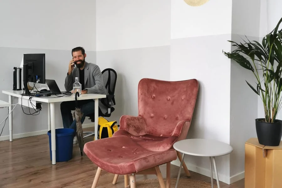 Ein Mann sitzt am Schreibtisch und telefoniert in einem modernen Büro mit einem Computer, einem rosa Sessel, einem Beistelltisch und einer Topfpflanze.