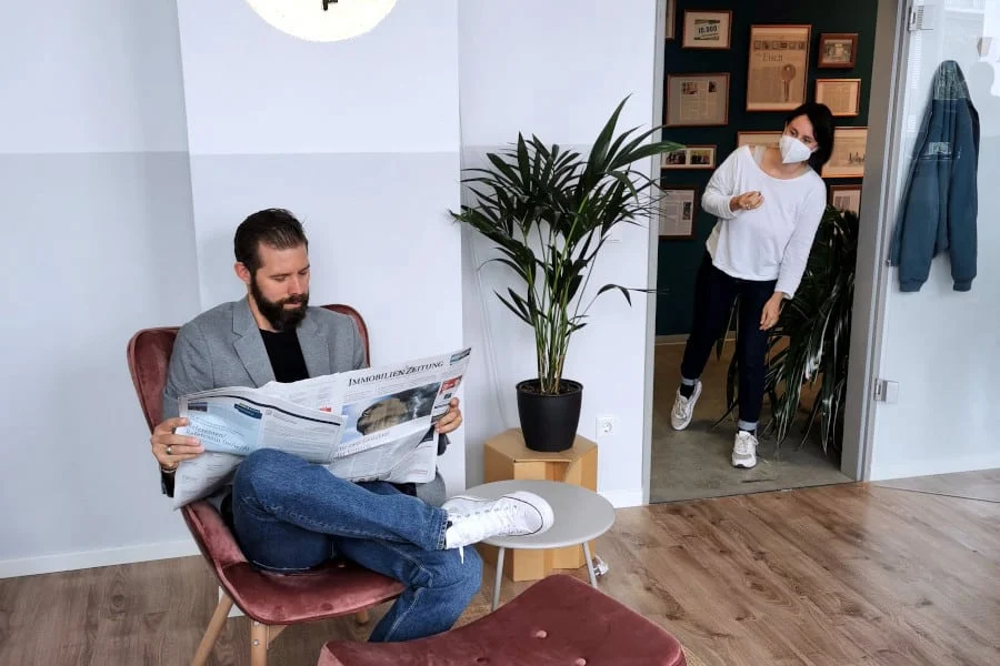 Ein Mann sitzt auf einem Stuhl und liest eine Zeitung, während eine Frau mit einer Maske und einem Gebäckstück den Raum betritt.