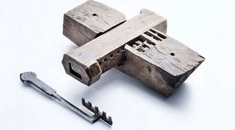 Ein großes, rechteckiges, hölzernes Schloss mit einem komplexen inneren Mechanismus wird neben einem altmodischen Metallschlüssel gezeigt.