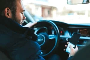 Ein Mann sitzt auf dem Fahrersitz eines Autos, hält mit einer Hand das Lenkrad fest und schaut mit der anderen Hand auf sein Smartphone.