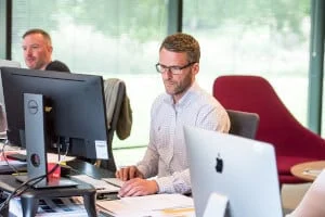 Zwei Männer arbeiten an Schreibtischen mit Computern in einem modernen, hell erleuchteten Büroraum mit großen Fenstern und einem roten Stuhl im Hintergrund.