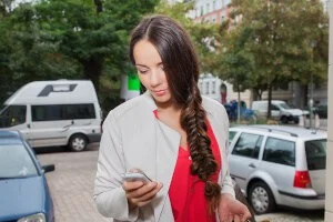 Eine Frau mit langen braunen Haaren, die zu einem Zopf geflochten sind, schaut auf ihr Smartphone, während sie im Freien in der Nähe von geparkten Autos auf einer Stadtstraße steht.