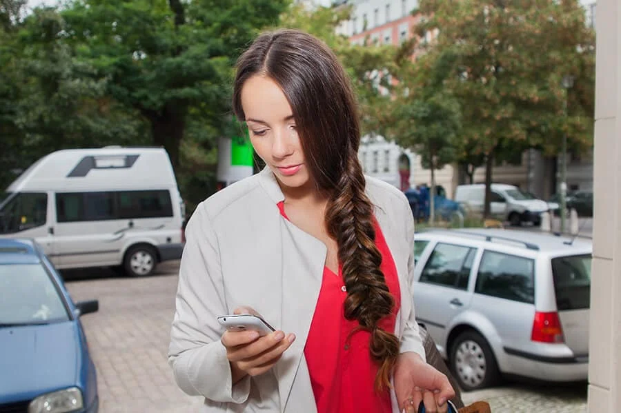 Eine Frau mit langen braunen Haaren, die zu einem Zopf geflochten sind, schaut auf ihr Smartphone, während sie im Freien in der Nähe von geparkten Autos auf einer Stadtstraße steht.