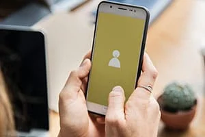 Person, die ein Smartphone mit einem gelben Bildschirm hält, der ein einfaches Benutzerprofilsymbol anzeigt, an einem Schreibtisch sitzend, mit einem Laptop und einer kleinen Topfpflanze im Hintergrund.