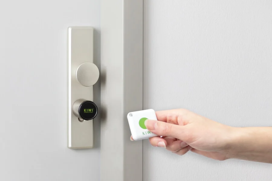 Eine Hand hält einen weißen elektronischen Schlüsselanhänger neben einem intelligenten Türschloss mit der Aufschrift "KIWI" an einer hellen Tür.