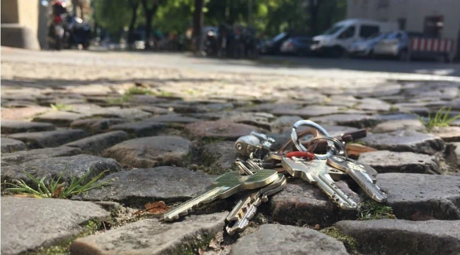 Ein Schlüsselbund liegt auf einem Kopfsteinpflaster im Freien, im Hintergrund sind Autos, Bäume und Gebäude zu sehen.