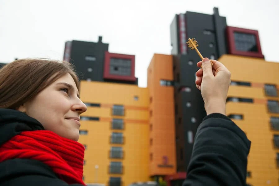 Eine Frau mit rotem Schal hält einen Schlüssel vor einem modernen Wohnhaus mit orangefarbenen und schwarzen Außenwänden hoch.