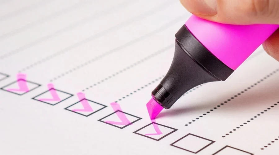 Eine Hand benutzt einen rosafarbenen Textmarker, um Kästchen auf einer Papier-Checkliste anzukreuzen und mehrere erledigte Aufgaben zu markieren.