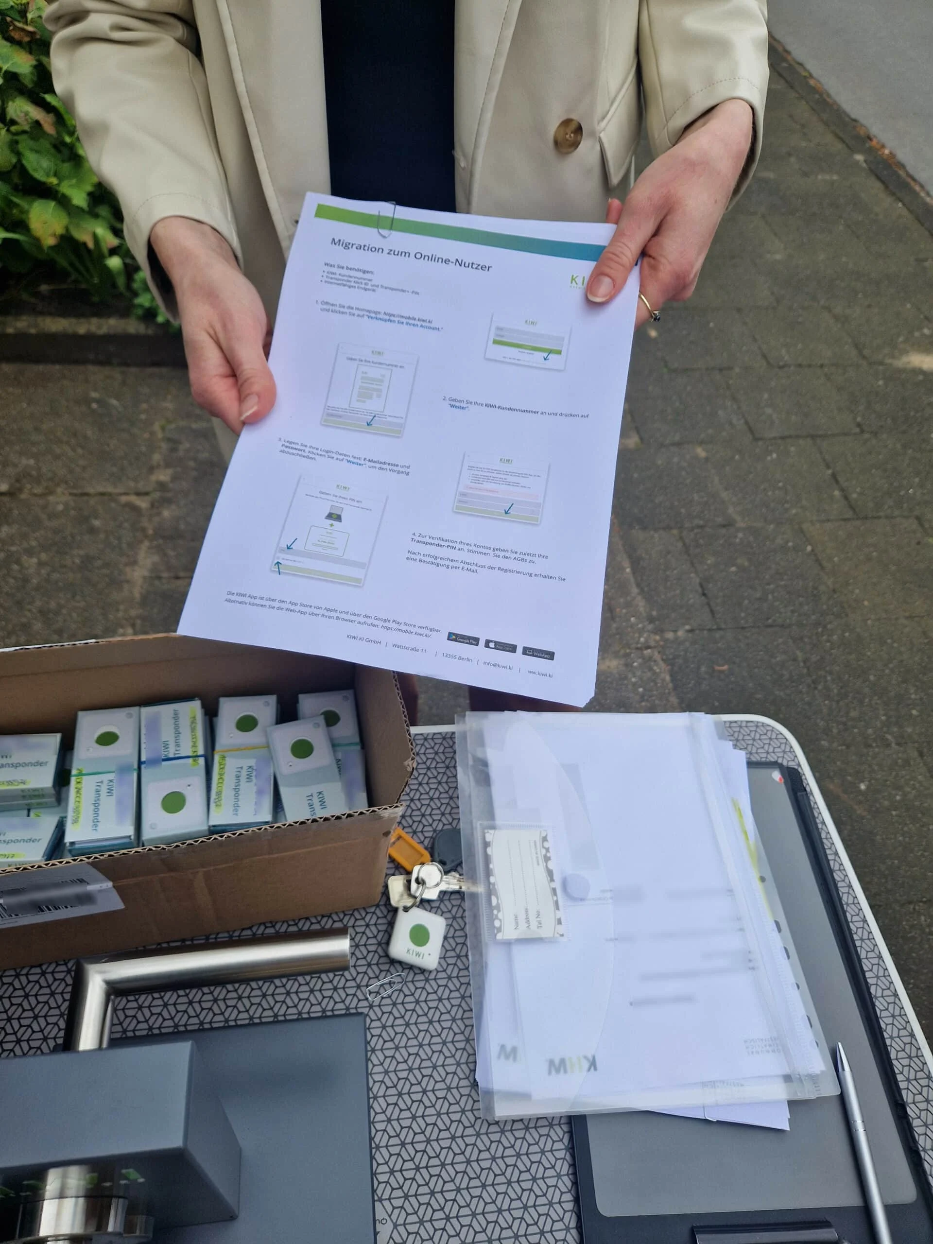Eine Person hält ein Anleitungsblatt neben einer Schachtel mit elektronischen Bauteilen, Papierkram und einem Schlüsselbund auf einem Tisch im Freien.