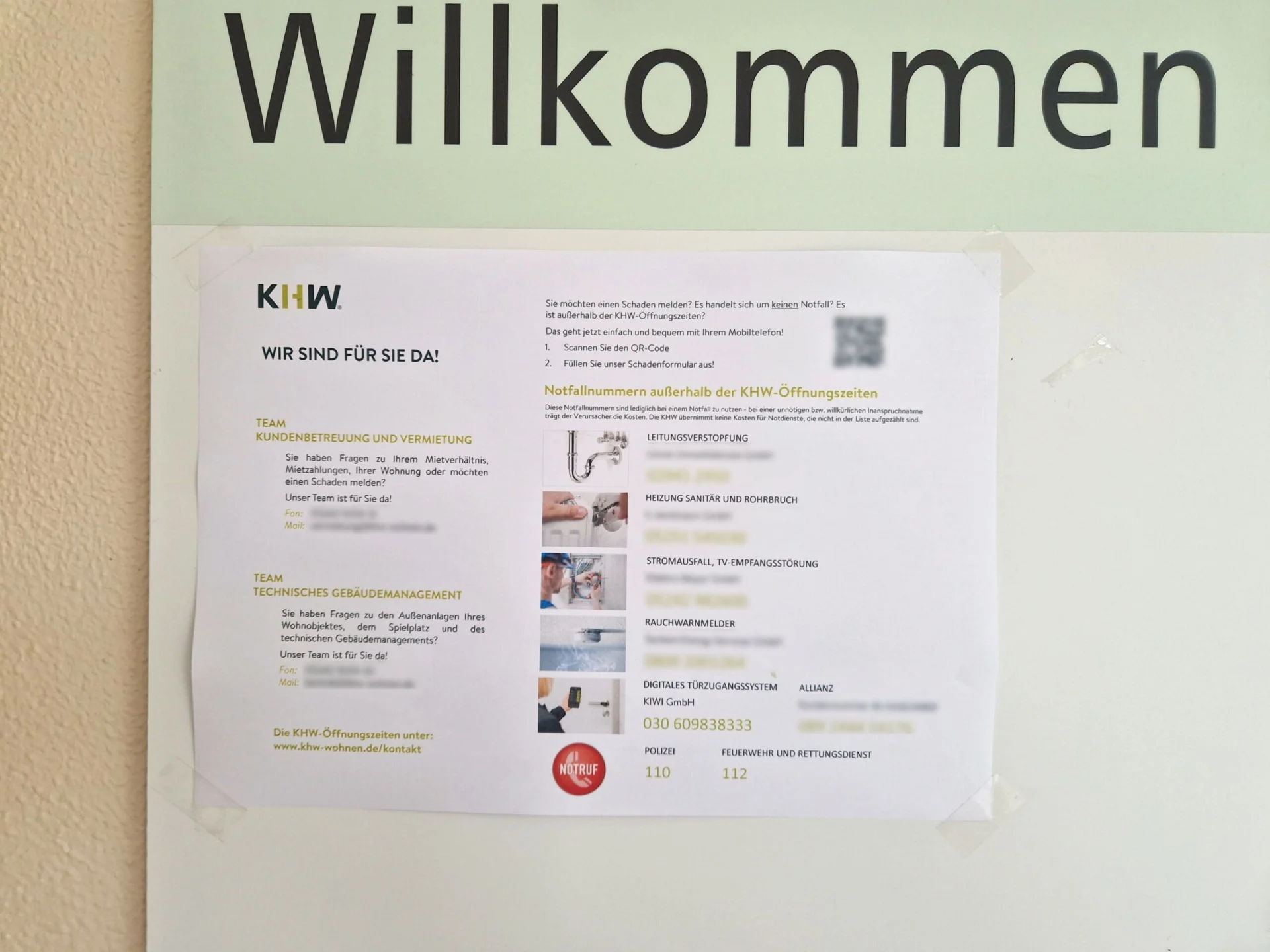 Ein gedruckter Zettel mit Kontaktinformationen und Notrufnummern in deutscher Sprache ist an einer Wand unter einem Schild mit der Aufschrift "Willkommen" angebracht.