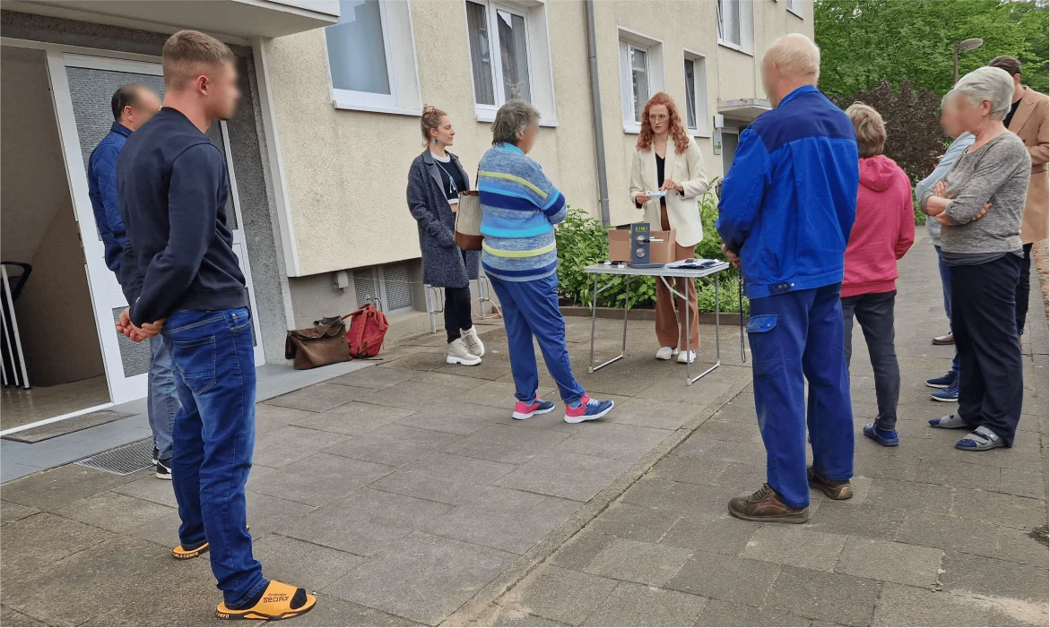 Eine Gruppe von Personen steht vor einem Gebäude in der Nähe eines Tisches, an dem eine Frau spricht oder einen Vortrag hält. Mehrere Zuhörer hören aufmerksam zu.