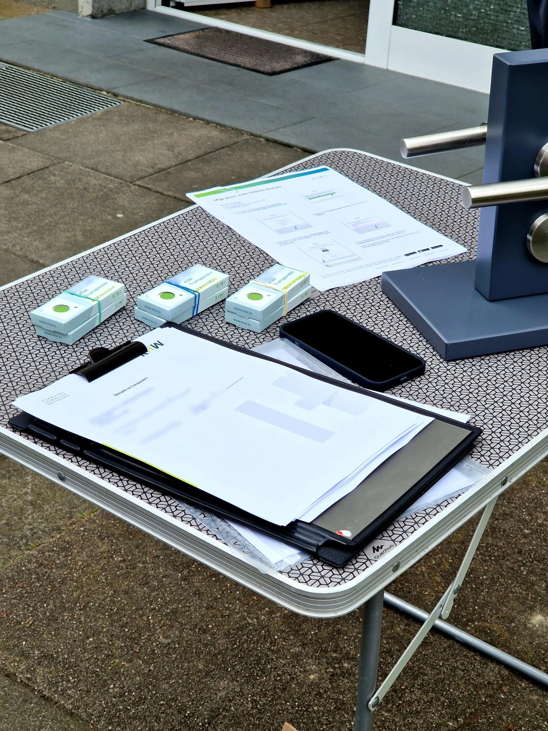 Ein Tisch mit Papierkram, kleinen Schachteln, einem Smartphone und einem Türgriff-Demonstrationsmodell, das im Freien auf einer gemusterten Fläche aufgestellt ist.