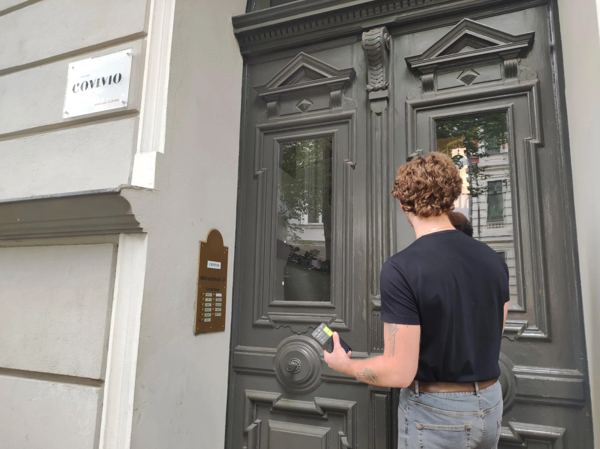 Eine Person mit kurzen lockigen Haaren und schwarzem T-Shirt steht an einer grauen Tür, benutzt eine Sprechanlage und hält ein kleines Gerät in einer Hand. Auf einem Schild an der Wand steht "CONVIVO".