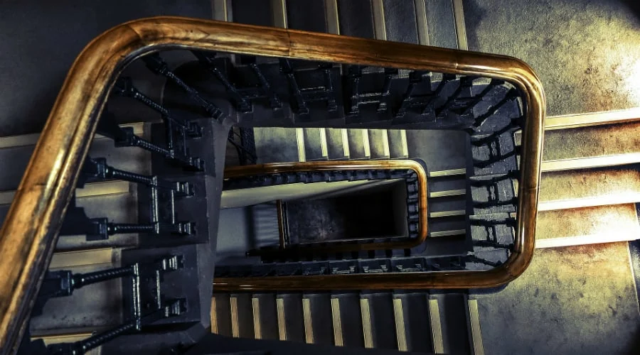 Blick von oben auf eine Wendeltreppe mit hölzernem Handlauf, schwarzen Metallgeländerstäben und beigen Stufen, die in einem quadratischen Muster nach unten führen.